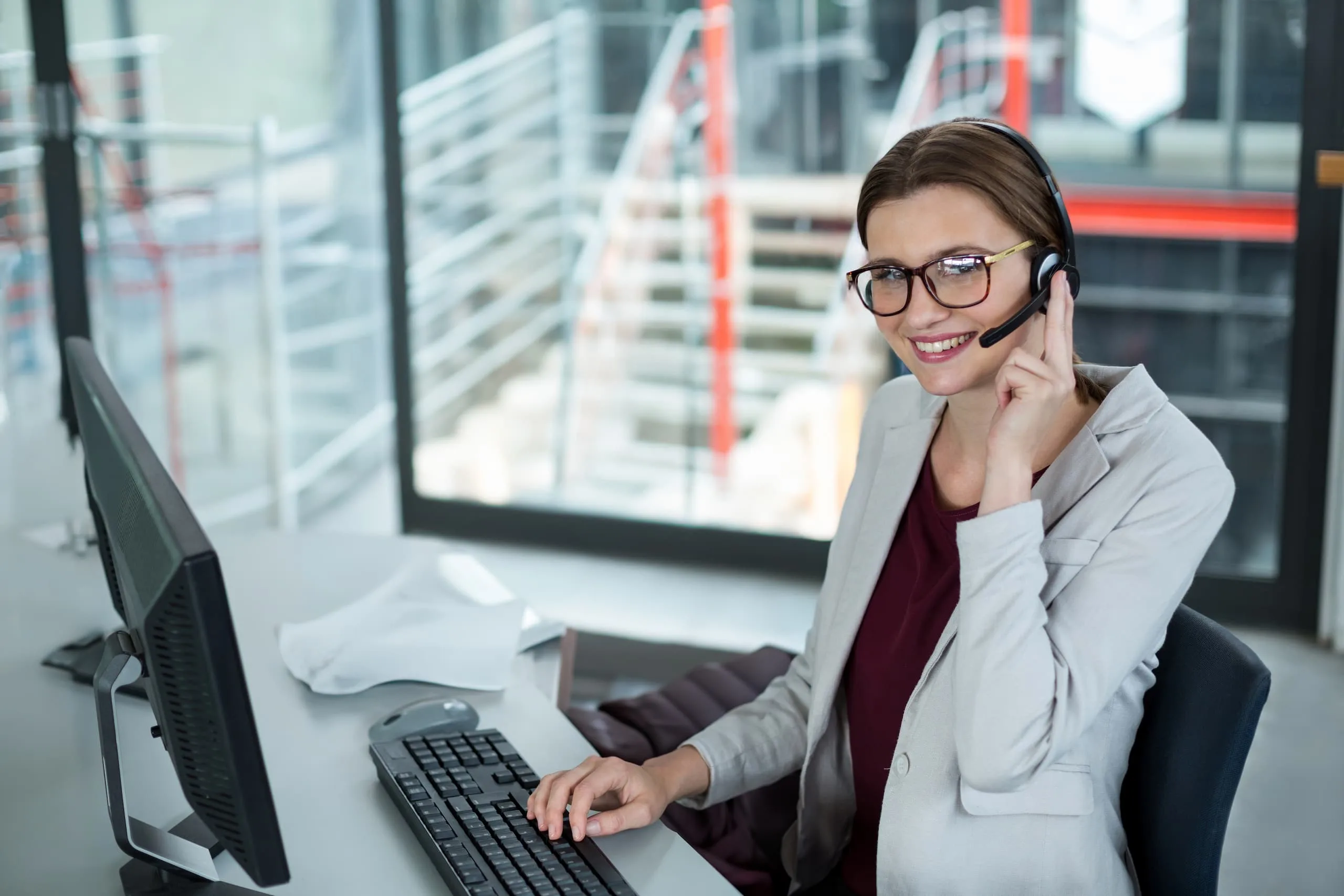 Eine lächelnde junge Frau mit Headset und Brille sitzt an einem Schreibtisch vor einem Computer und arbeitet.