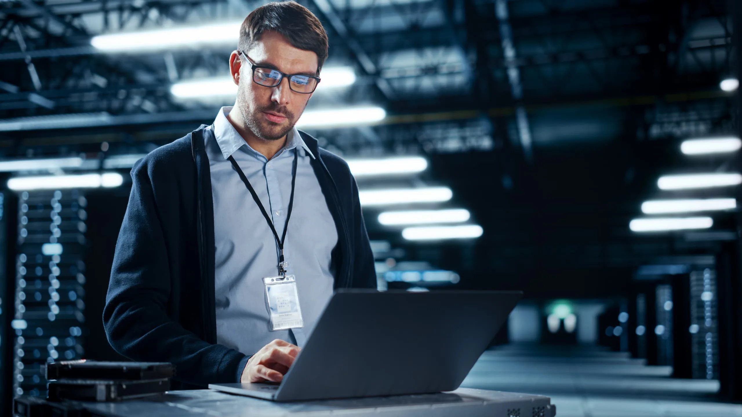 Ein Mann mit Brille und Ausweis steht in einem Serverraum und arbeitet an einem Laptop.